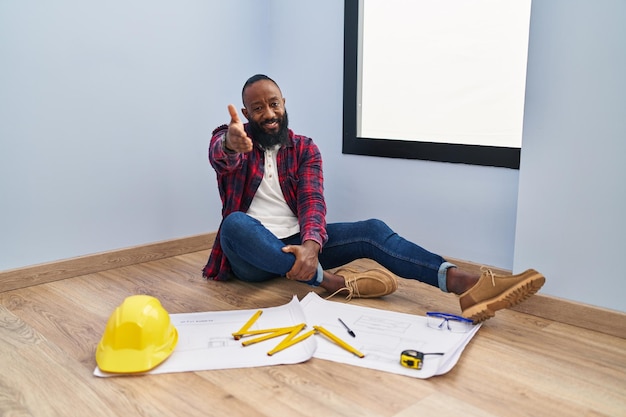 African american man sitting on the floor at new home looking at blueprints smiling friendly offering handshake as greeting and welcoming successful business