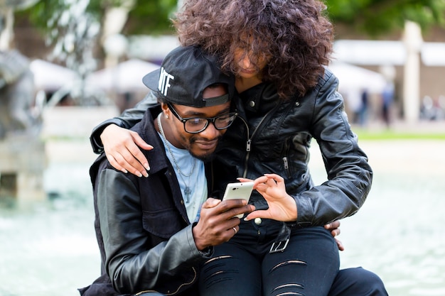 Photo african american man sitting with girlfriend at fountain showing message on mobile phone or looking up information