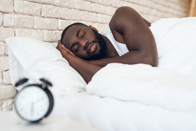 Афро-американский мужчина спит в постели рядом с будильником