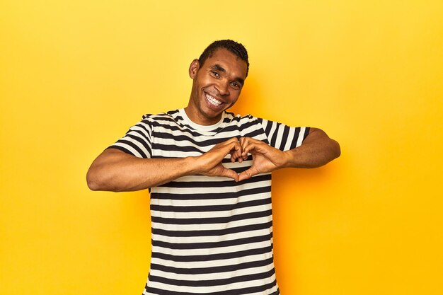 African american man in striped tshirt yellow studio smiling and showing a heart shape with hands