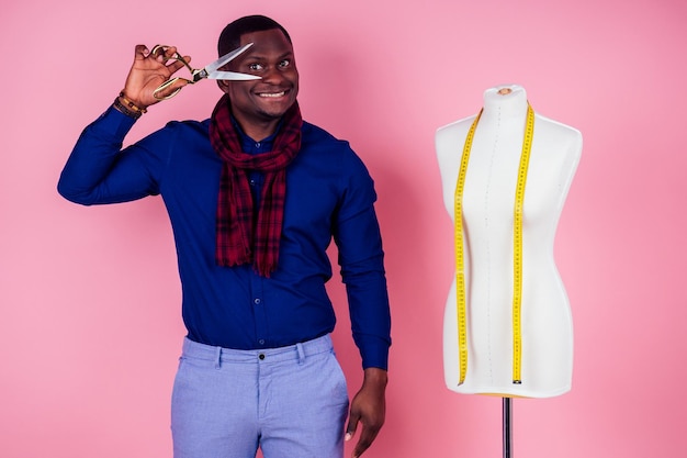 African American man tailor seamstress workshop stylish male model clothes designer measuring tape on neck posing next to the mannequin holding scissors in hand on pink background in the studio