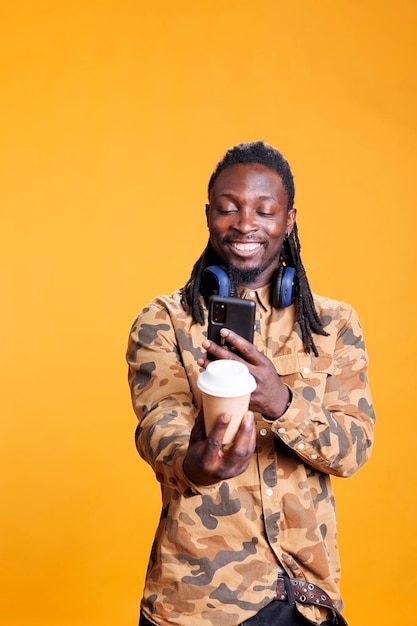 African american man taking photo of cup with coffee using mobile phone, enjoying free time in studio over yellow background. cheerful smiling person drinking beverage, listening music and having fun