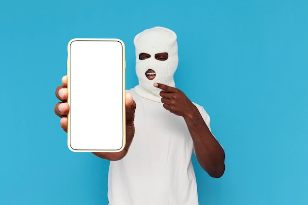 african american man thug in white balaclava shows empty smartphone screen on blue background