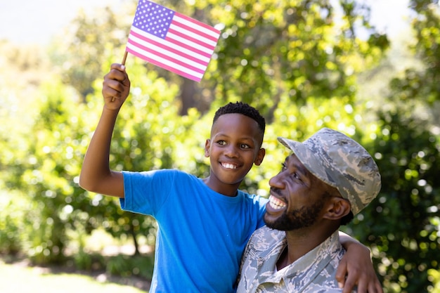 軍服を着たアフリカ系アメリカ人男性が家に帰り、息子を抱き上げ、カメラを見て微笑み、晴れた日に少年がミニ旗を持っている