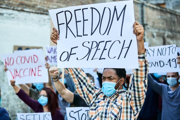 African American man wearing protective face mask while participating in public demonstrations and carrying banner with 'freedom of speech' inscription