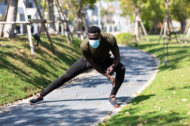 African american man wearing protective medical face mask\
jogging and running beside road in park at morning.