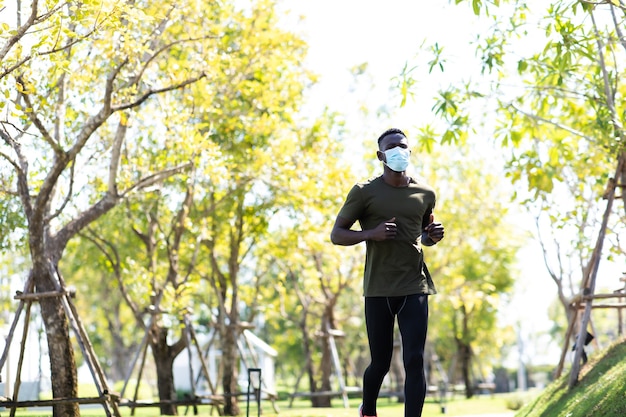 African american man wearing protective medical face mask\
jogging and running beside road in park at morning.