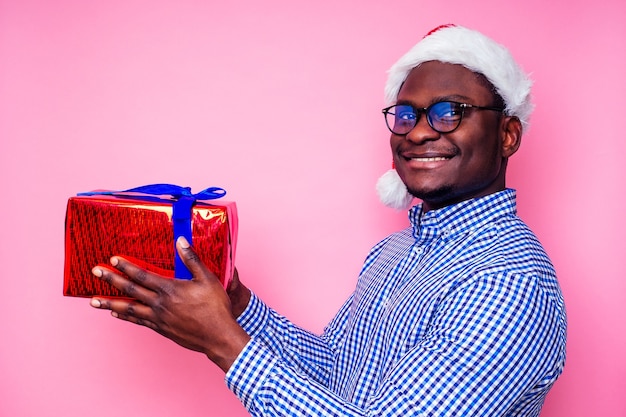 세련된 격자 무늬 셔츠를 입은 아프리카계 미국인 남자는 분홍색 배경 스튜디오에 선물 상자가 있는 산타 모자에 큰 미소를 지으며 메리 크리스마스를 축하하는 어두운 피부의 산타클로스