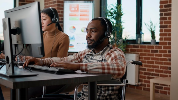 African american man in wheelchair answering helpline call at customer service job, working in disability friendly office. Male worker using telework headset at helpdesk to help clients.