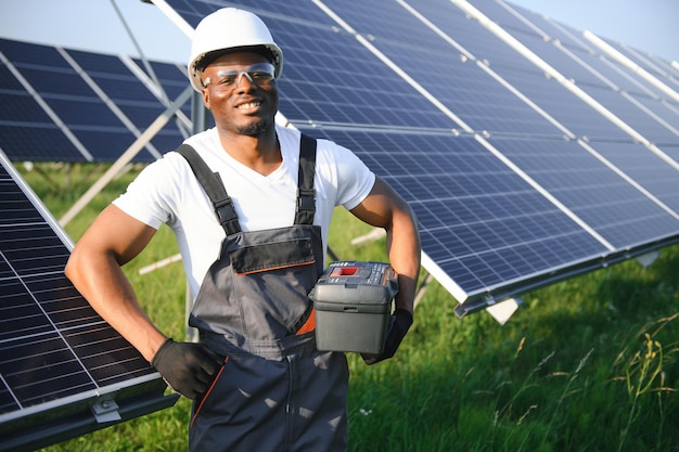 African american man in white helmet and grey overalls standing among rows of solar panels