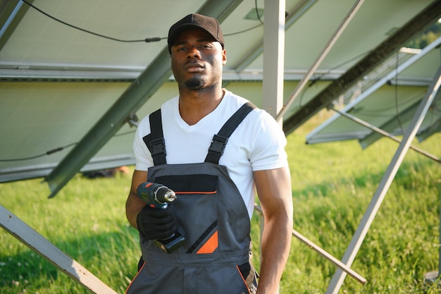 African american man in white helmet and grey overalls standing among rows of solar panels