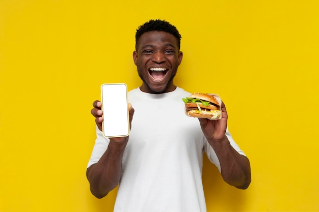 African american man in white tshirt holds big burger and shows blank smartphone screen