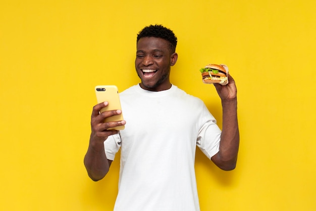 African american man in white tshirt holds big burger and uses smartphone on yellow background