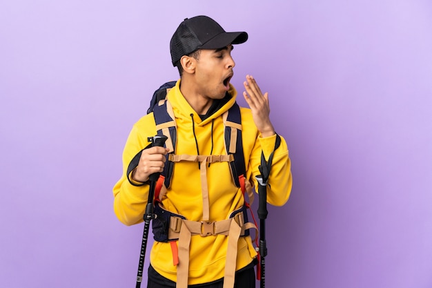 Фото Афро-американский мужчина с рюкзаком и треккинговыми палками над изолированной стеной, зевая и прикрывая широко открытый рот рукой