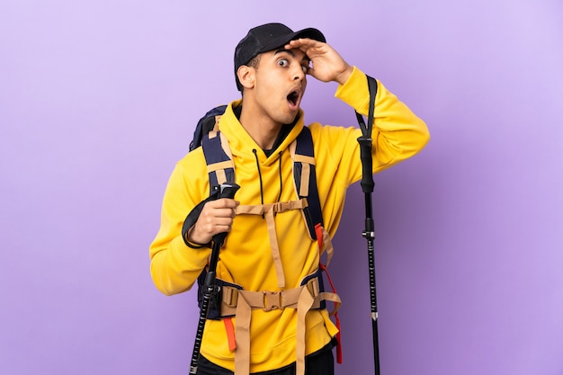 African American man with backpack and trekking poles over isolated wall doing surprise gesture while looking to the side