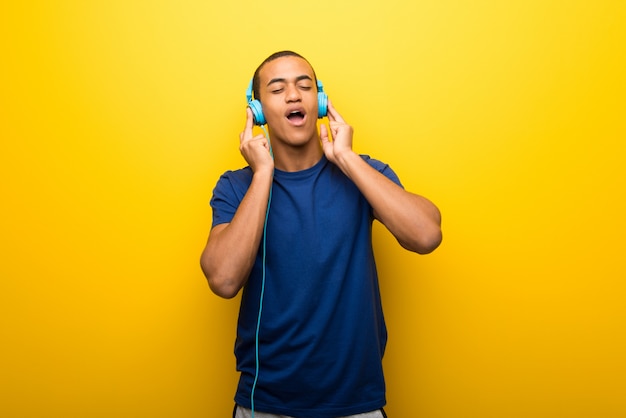 ヘッドフォンで音楽を聴く黄色の壁に青いtシャツを持つアフリカ系アメリカ人