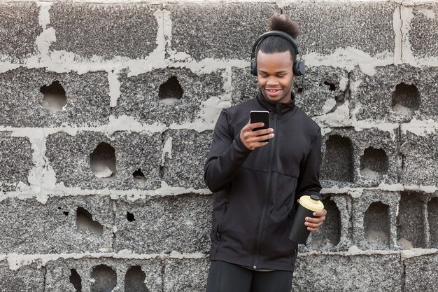 African American man with drink using smartphone near wall