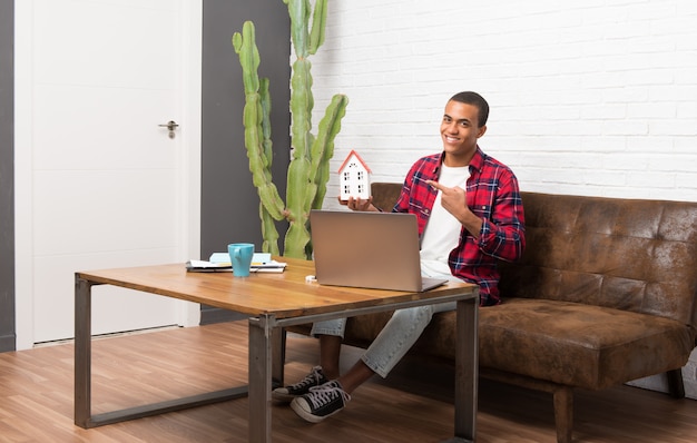 African american man with laptop in the living room holding a little house