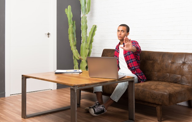 L'uomo afroamericano con il computer portatile nel soggiorno che fa il gesto di arresto che nega una situazione che pensa sbagliato