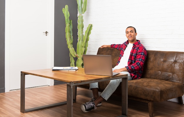 African american man with laptop in the living room posing with arms at hip and smiling