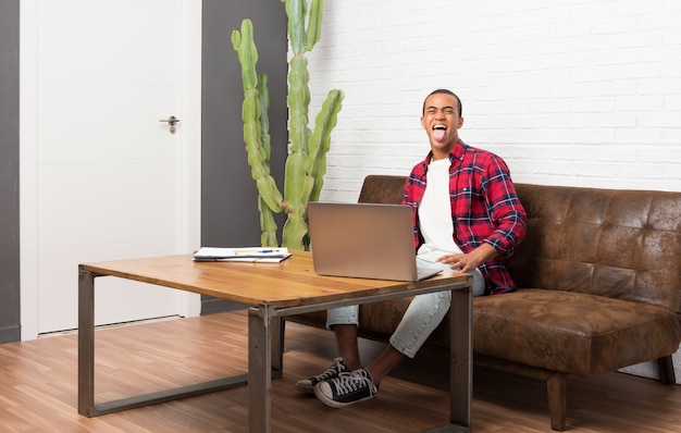 Foto uomo afroamericano con il computer portatile nel salone che mostra lingua alla macchina fotografica che ha sguardo divertente