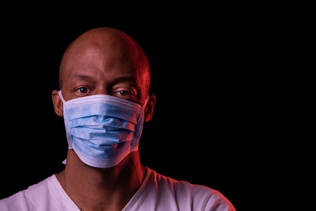 Foto uomo afroamericano con la maschera protettiva del coronavirus che esamina la macchina fotografica su fondo scuro