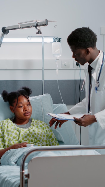 African american man and woman consulting patient in bed to treat sickness. Medical team of specialists helping young adult with IV drip bag and heart rate monitor in hospital ward