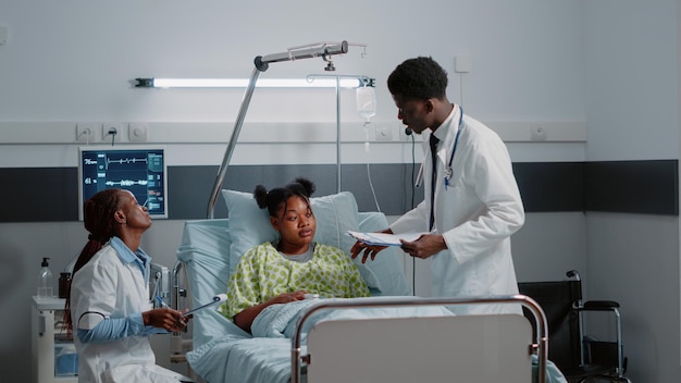 African american man and woman consulting patient in bed to treat sickness. Medical team of specialists helping young adult with IV drip bag and heart rate monitor in hospital ward