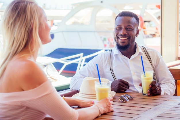 african american man and a woman look lovingly at each in summer tropical cafe and drinking fresh fruit orange cocktail.