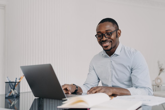 オンラインでラップトップに取り組んでいるアフリカ系アメリカ人の男性は、彼のビジネスプロジェクトの良い仕事の笑顔に満足しています