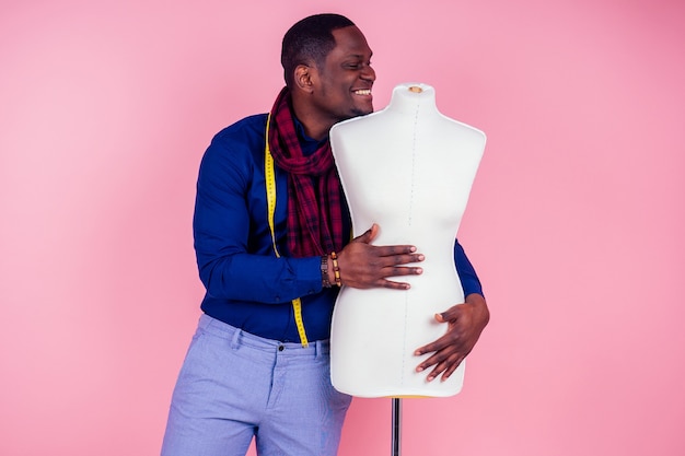 African American man working in a tailor seamstress workshop stylish male model clothes designer with a measuring tape on neck posing next to the mannequin on pink background in the studio