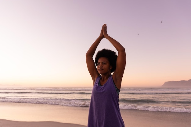 Photo african american mature woman with arms raised exercising at beach against clear sky during sunset