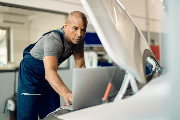 African American mechanic using laptop while examining car engine in repair shop