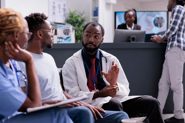 Equipe medica afroamericana che consulta l'uomo seduto nella sala d'attesa nella hall della reception della struttura. medico e assistente che parlano con il paziente della diagnosi e del trattamento della malattia, supporto sanitario.