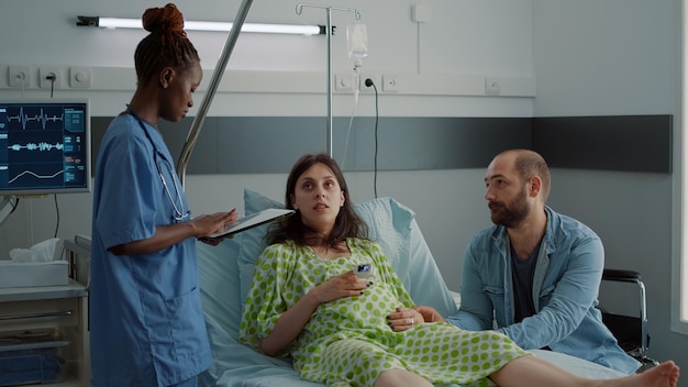 Foto infermiera afroamericana che utilizza tablet per l'assistenza sanitaria delle pazienti in gravidanza in corsia ospedaliera. donna in attesa con pancione seduto con l'uomo alla maternità. coppia con specialista in ostetricia