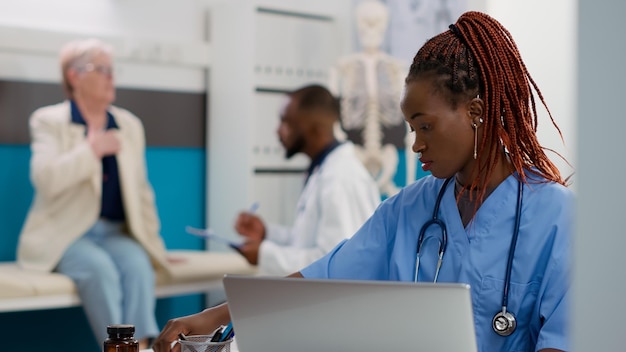 African american nurse working on appointments in cabinet,\
using laptop to plan checkup visits to help patients at healthcare\
facility. medical assistant with stethoscope planning\
consultations.