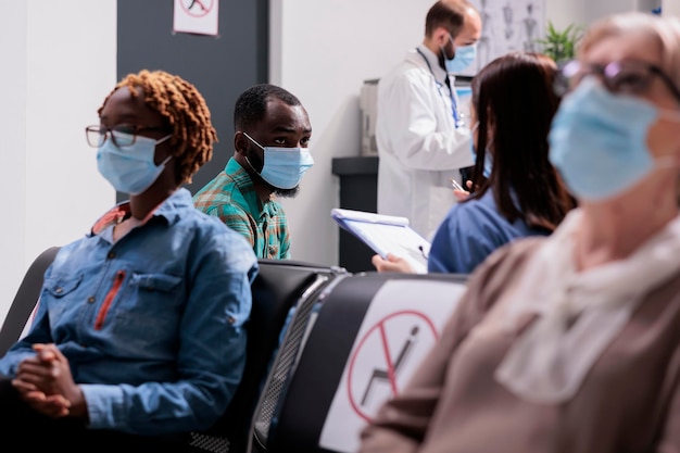 African american patient attended by female doctor. Different ethnicities, ages people wearing masks preventing covid 19 spread at doctor office reception area, general shot of hospital waiting room.