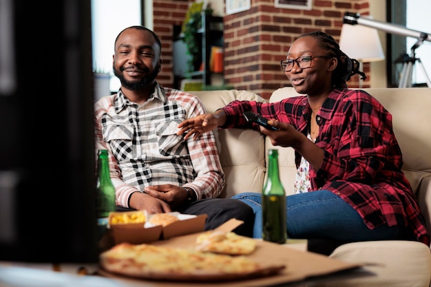 African american people switching tv channels and eating\
takeaway meal from fast food delivery at home. watching movie on\
television program and enjoying takeout food with beer\
bottles.