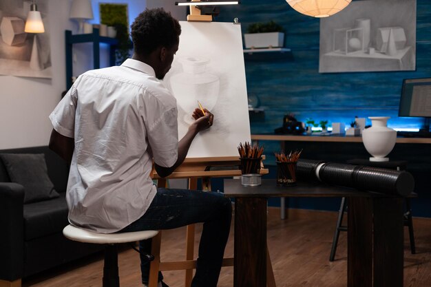 African american person with artistic abilities putting\
finishing touches on pencil drawing of vase. talented young man\
creating pot outline on canvas easel in home crafts atelier.