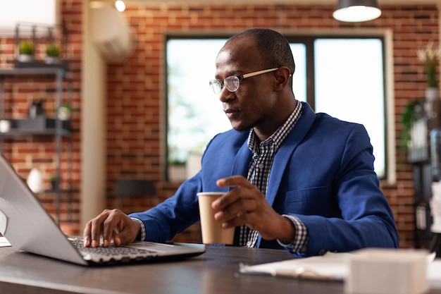 Афроамериканец работает над бизнес-проектом с ноутбуком и держит чашку кофе. Сотрудник работает на компьютере для анализа лидерства и маркетинговой стратегии. Мужчина на работе в компании