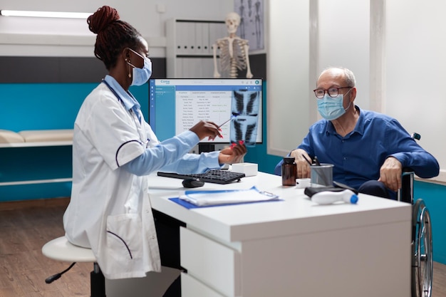 African american physician doctor with face mask against covid19 explaining radiography expertise to sick senior man during medical appointment in hospital office. Medicine concept