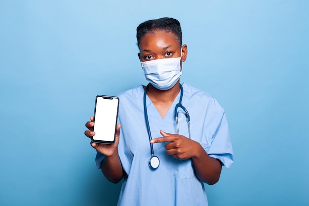 Infermiere medico afroamericano che indossa una maschera protettiva per prevenire l'infezione da coronavirus che tiene in mano uno smartphone con schermo bianco che lavora in studio con sfondo blu. concetto di medicina