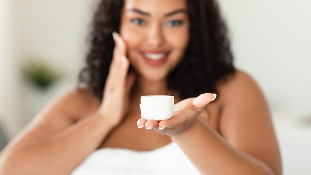 African american plus size woman holding jar of skin cream applying moisturiser on face after shower selective focus