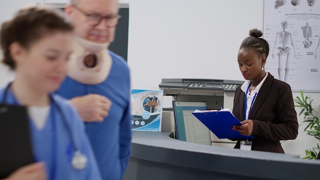 African american receptionist helping patient with neck collar\
at hospital reception counter, nurse taking old man with cervical\
foam. having medical appointment checkup visit, facility\
lobby.