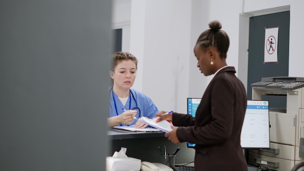 Photo african american receptionist working with nurse at reception counter, making registration appointments with checkup reports. diverse team talking about healthcare insurance and medical forms.