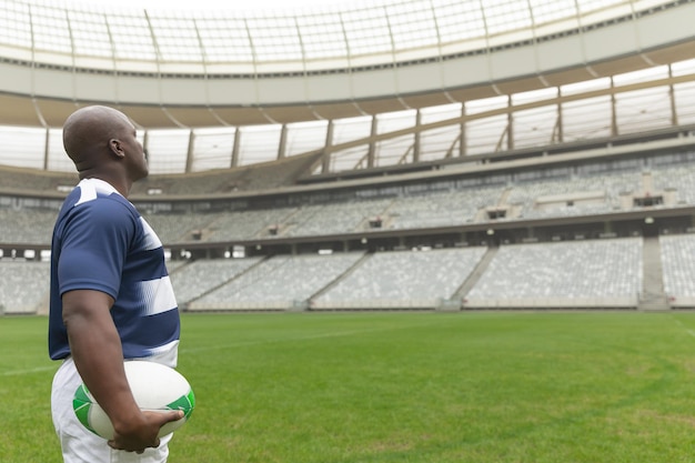 African american rugby player standing with rugby ball and looking away in stadium