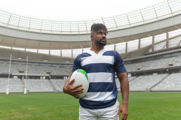 african american rugby player standing with rugby ball and looking away in stadium