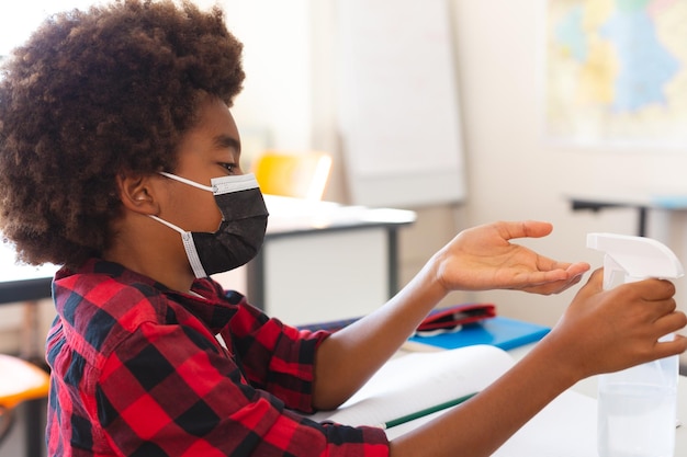 African american schoolboy in face mask sitting at desk in classroom disinfecting hands