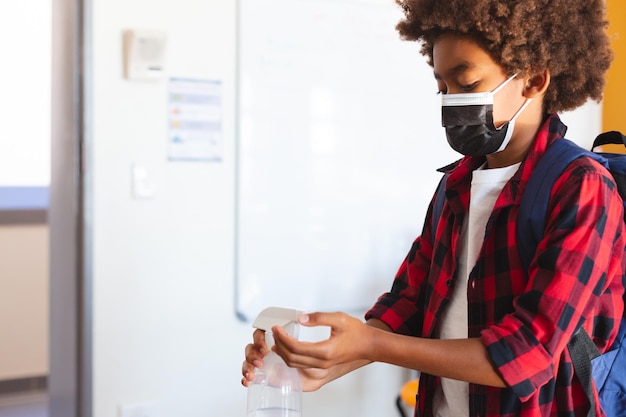 African american schoolboy in face mask standing in classroom disinfecting hands