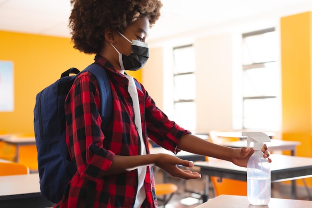 African american schoolboy in face mask standing in classroom disinfecting hands